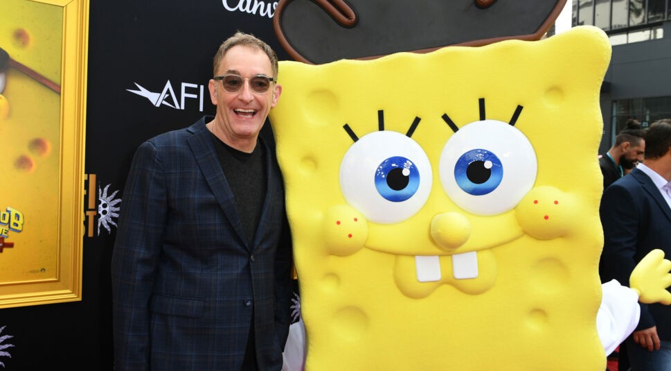 Auf dem Bild steht Tom Kenny, der englische SpongeBob-Synchronsprecher, lachend neben einer lebensgroßen SpongeBob-Figur in Piratenverkleidung. Tom Kenny trägt ein kariertes Sakko und eine Sonnenbrille. Die Szene spielt sich auf einem roten Teppich bei einer Filmpremiere ab. Die Stimmung ist ausgelassen und fröhlich.