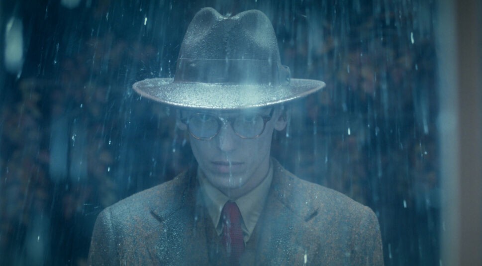 Bei Stranger Things Staffel 5 siehst du Henry Creel, gespielt von Jamie Campbell Bower, in einer düsteren Regenszene. Er trägt einen nassen Fedora-Hut und eine altmodische Brille, sein Blick ist ernst und geheimnisvoll.