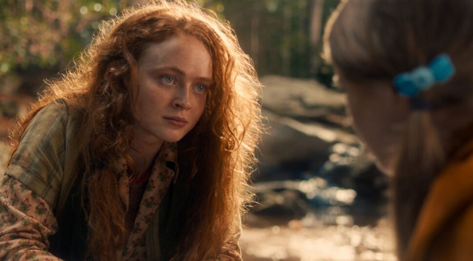 Bei Stranger Things Staffel 5 siehst du Max Mayfield, gespielt von Sadie Sink, in einem ruhigen Moment im Freien. Mit ernstem Blick und zerzausten roten Locken kniet sie vor einem Kind mit blauer Haarspange.