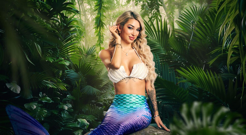 Auf dem Bild zum Dschungelcamp 2026 ist Teilnehmerin Ariel in einer fantasievollen Meerjungfrauen-Verkleidung zu sehen. Sie sitzt auf einem Felsen im Dschungel, trägt ein Muschel-Bikini-Top und eine schimmernde Fischschwanzflosse in Blau-Lila. Mit lockigem, blondem Haar und einem sanften Lächeln wirkt sie verspielt und selbstbewusst vor tropischem Hintergrund.