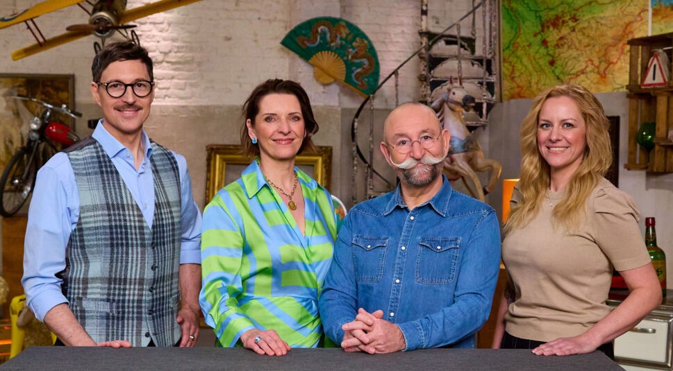 Auf dem Bild mit den Bares für Rares-Expert:innen stehen vier bekannte Gesichter der Trödelshow nebeneinander vor einem Studiotresen. Von links nach rechts sind Colmar Schulte-Goltz mit kariertem Anzug, Wendela Horz in einer blau-grünen Bluse, Moderator Horst Lichter mit rundem Brillenrahmen und Schnurrbart sowie Annika Raßbach in einem beigefarbenen Oberteil zu sehen. Die Atmosphäre wirkt offen und herzlich.