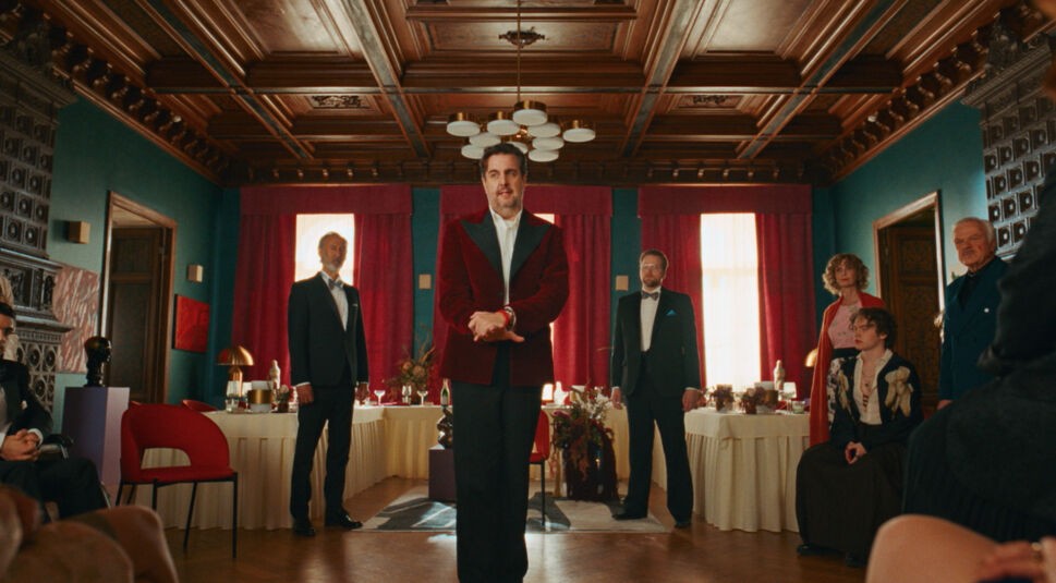 Auf dem Bild in dem Artikel zu "Fabian und Die mörderische Hochzeit" steht eine Gruppe elegant gekleideter Menschen in einem Saal mit Holzdecke und roten Vorhängen.