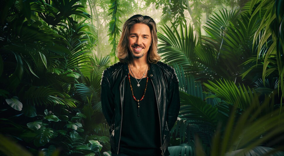 Auf dem Bild zum Dschungelcamp 2026 steht Teilnehmer Gil Ofarim vor einem dichten, grünen Dschungelhintergrund. Er trägt eine schwarze Lederjacke über einem dunklen Shirt und mehrere auffällige Ketten, darunter eine mit Davidstern. Mit schulterlangem Haar, lässigem Lächeln und charismatischer Ausstrahlung wirkt er entspannt, aber kampfbereit für das Abenteuer.