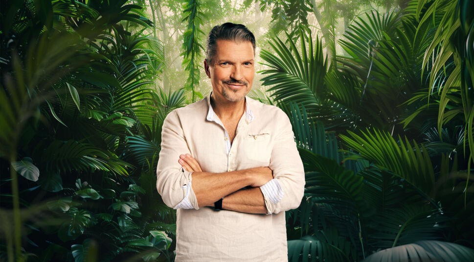 Teilnehmer Hardy Krüger steht auf dem Bild zum Dschungelcamp 2026 vor einer grünen, tropischen Dschungelkulisse. Er trägt ein beigefarbenes Leinenhemd mit aufgekrempelten Ärmeln und verschränkten Armen. Mit graumelierten Haaren, dezentem Bart und einem ruhigen Lächeln wirkt er souverän, erfahren und entspannt vor dem Start ins Dschungelabenteuer.