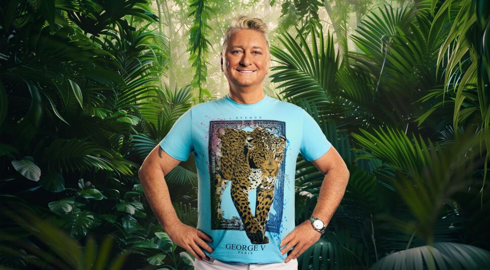 Auf dem Bild zum Dschungelcamp 2026 steht Teilnehmer Hubert Fella vor dichtem, tropischem Dschungelgrün. Er trägt ein auffälliges, hellblaues T-Shirt mit einem großen Gepardenmotiv der Marke George V Paris und kombiniert es mit einer weißen Hose. Mit beiden Händen in der Hüfte und einem breiten Lächeln zeigt er sich farbenfroh, offen und gut gelaunt.