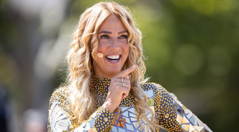 Auf dem Bild zu Kampf der RealityAllstars 2026 ist die Sängerin und Reality-TV-Teilnehmerin Loona fröhlich lächelnd zu sehen. Sie trägt ein auffälliges Glitzeroutfit mit goldenen und silbernen Spiegelelementen. Mit einem Headset-Mikrofon ausgestattet, zeigt sie mit dem Finger nach vorn. Die Stimmung ist gelöst und positiv, das Bild wurde im Freien aufgenommen.