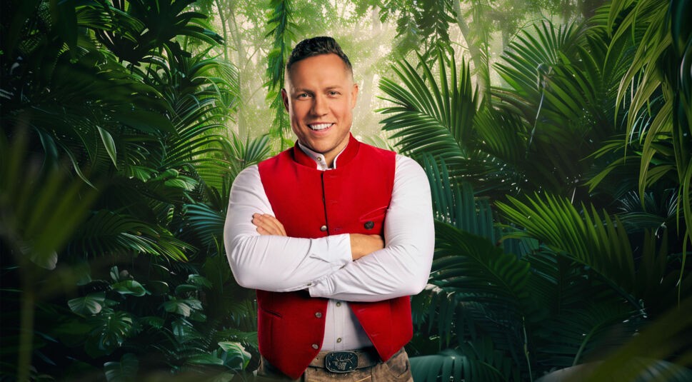 Teilnehmer Patrick Romer steht auf dem Bild zum Dschungelcamp 2026 selbstsicher vor dichtem, grünem Dschungelhintergrund. Er trägt ein weißes Hemd mit einer traditionellen roten Trachtenweste und einer Gürtelschnalle mit Hirschmotiv. Mit verschränkten Armen und breitem Lächeln vermittelt er eine bodenständige, zugleich entschlossene Ausstrahlung.
