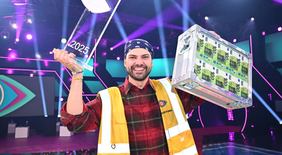 Auf dem Bild aus Promi Big Brother strahlt Jimi Blue Ochsenknecht als Gewinner der Staffel 2025. Er trägt eine gelbe Warnweste mit der Aufschrift "Projektleitung", hält eine durchsichtige Trophäe mit der Jahreszahl 2025 in der linken Hand und einen silbernen Geldkoffer voller 100-Euro-Scheine in der rechten Hand. Die Bühne ist bunt beleuchtet, die Stimmung ist feierlich und ausgelassen.