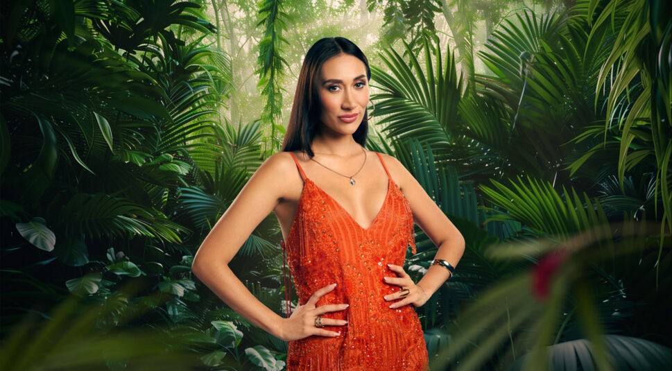 Auf dem Bild zum Dschungelcamp 2026 posiert Teilnehmerin Samira Yavuz vor tropischem Blätterdschungel. Sie trägt ein auffälliges, tief ausgeschnittenes Glitzerkleid in leuchtendem Orange mit funkelnden Details. Ihre langen, dunklen Haare fallen offen über die Schultern. Mit Händen in der Taille und selbstbewusstem Blick wirkt sie glamourös und kampfbereit fürs Camp.