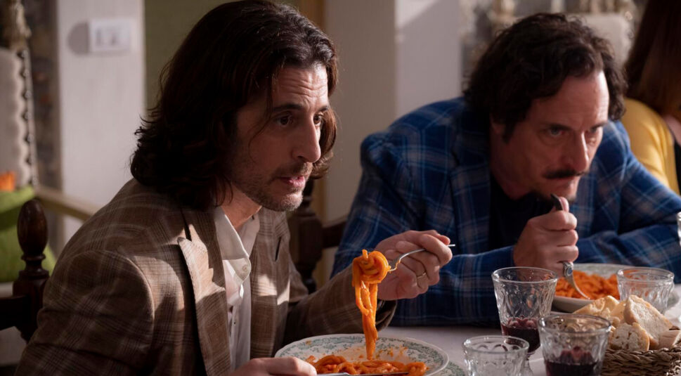 Auf dem Bild aus dem Film Solo Mio sitzen Neil, gespielt von Jonathan Roumie, und Julian, gespielt von Kim Coates, gemeinsam am Esstisch. Neil trägt ein kariertes Sakko und isst konzentriert Pasta, während Julian im blauen Flanellhemd ernst dreinblickt. Auf dem Tisch stehen Weingläser, Brot und Pasta – die Szene wirkt familiär und angespannt.