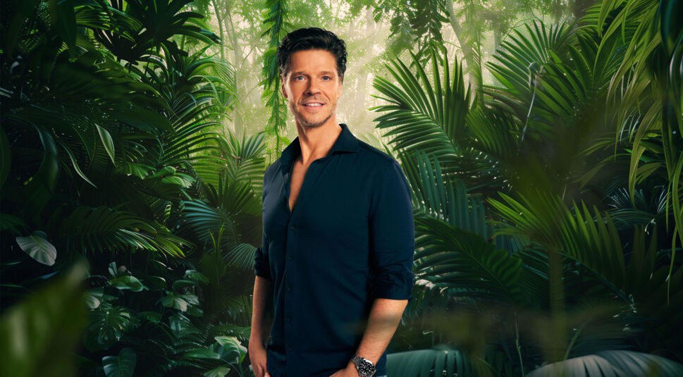 Auf dem Bild zum Dschungelcamp 2026 steht Teilnehmer Stephen Dürr locker und selbstbewusst vor einer grünen Dschungelkulisse. Er trägt ein dunkelblaues, aufgeknöpftes Hemd mit hochgekrempelten Ärmeln und eine Armbanduhr. Mit seinem offenen Blick, zurückgegeltem Haar und entspanntem Lächeln strahlt er Ruhe und Gelassenheit aus.