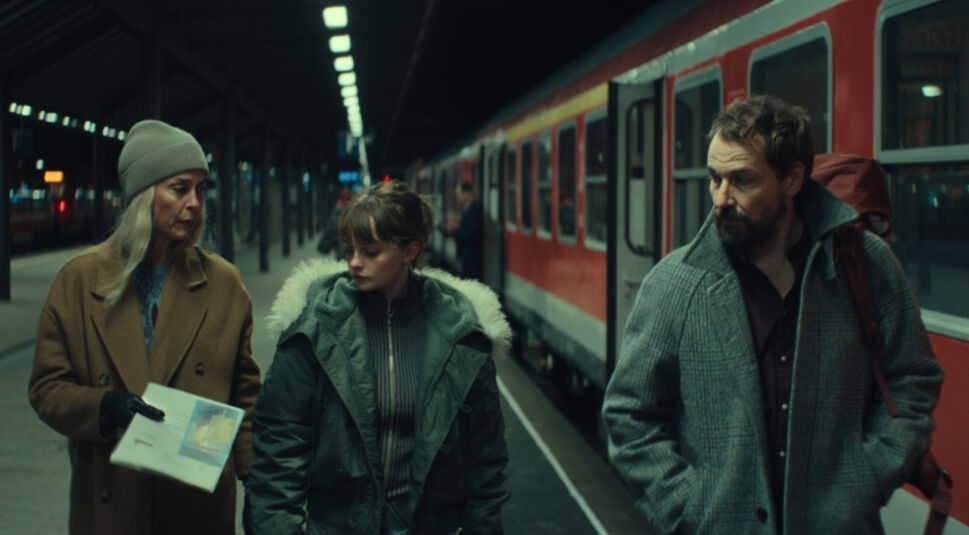 Meret und Simon gehen mit ihrer Tochter über einen Bahnhof und unterhalten sich dabe.