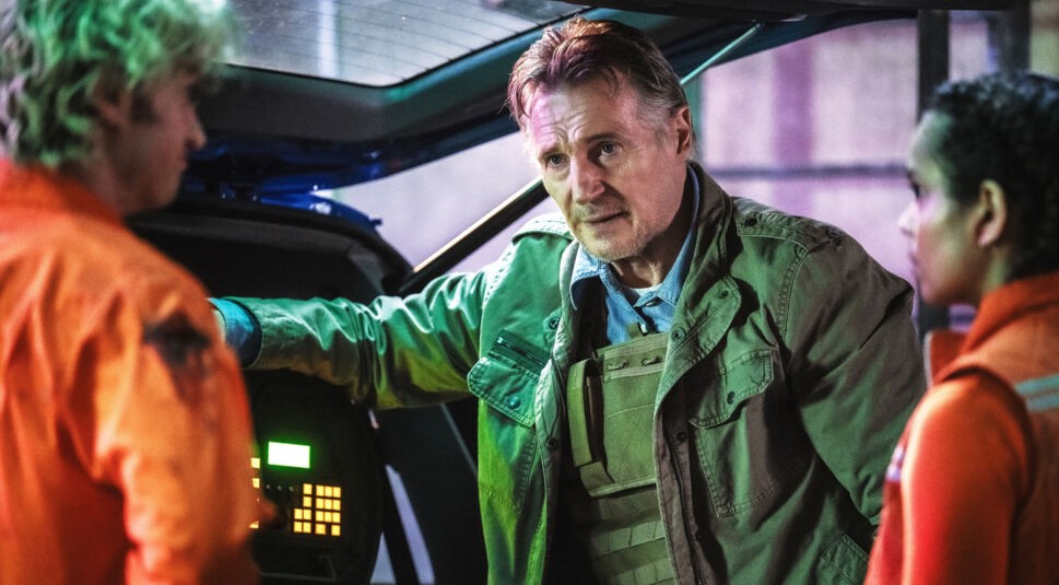 Auf dem Bild zur Cold Storage Filmkritik ist ein Bioterror-Agent zu sehen, gespielt von Liam Neeson, der ernst und kontrolliert vor zwei Personen in orangefarbener Schutzkleidung steht. In einer düsteren, technisch anmutenden Umgebung lehnt er an einem geöffneten Fahrzeug oder Gerät. Die grünliche Beleuchtung und seine angespannte Mimik vermitteln eine bedrohliche, konfliktreiche Stimmung.