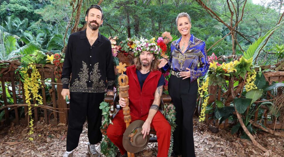 Auf dem Bild zum Dschungelcamp 2026 ist Gil als frisch gekrönter Dschungelkönig zu sehen. Er sitzt auf dem Thron, trägt die Krone und hält das Zepter in der Hand. Neben ihm stehen die Moderatorinnen Sonja und Jan, die stolz lächeln. Die Szene ist festlich, triumphal und von Dschungelkulisse umgeben.