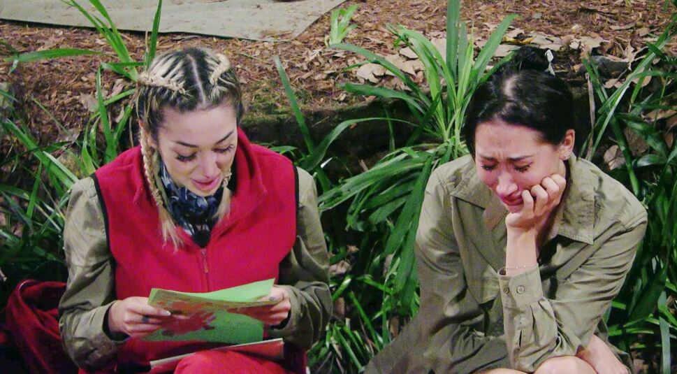 Auf dem Bild zum Dschungelcamp 2026 sind Ariel und Samira in einer emotionalen Szene zu sehen. Ariel sitzt links in roter Weste und liest konzentriert einen Brief, während Samira rechts daneben sichtlich gerührt wirkt und Tränen in den Augen hat. Umgeben von Dschungelpflanzen herrscht eine nachdenkliche, traurige Stimmung.