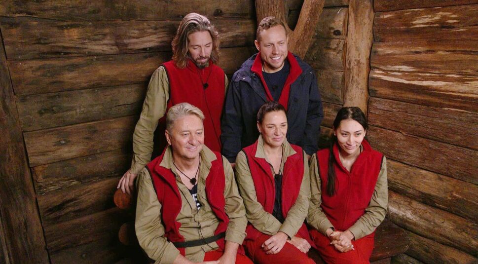 Auf dem Bild zum Dschungelcamp 2026 sind die Halbfinalisten Gil, Patrick, Hubert, Simone und Samira in einer gemeinsamen Szene zu sehen. Die Gruppe sitzt eng beieinander in einer hölzernen Dschungelprüfungskulisse und trägt rote Westen. Die Stimmung wirkt ruhig, angespannt und erwartungsvoll vor der nächsten Entscheidung.