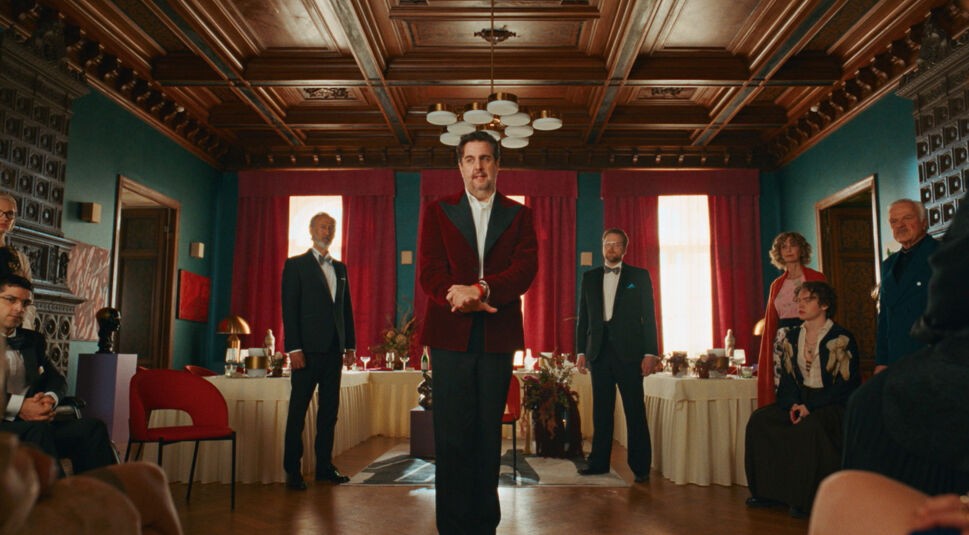 Auf dem Bild in dem Artikel zum Ende von "Fabian und Die mörderische Hochzeit" erklärt, ist der Hauptdarsteller in der Mitte eines Raumes zu sehen, umgeben von weiteren Charakteren.