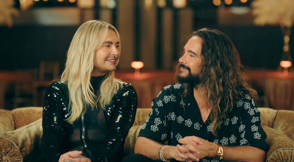 Auf dem Bild zu Kaulitz & Kaulitz Staffel 3 sitzen Bill und Tom Kaulitz gemeinsam auf einem Sofa. Bill trägt links ein schwarzes, glitzerndes Oberteil und hat langes blondes Haar. Tom sitzt rechts in einem gemusterten schwarzen Hemd, mit langem braunem Haar und Bart. Beide lächeln sich an, wodurch die Szene vertraut, herzlich und entspannt wirkt.