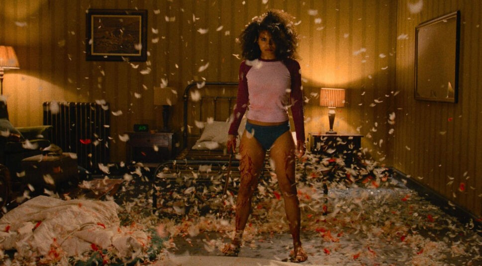 Zazie Beetz in They Will Kill You: Eine Frau steht blutverschmiert, mit einem Messer in der Hand in einem verwüsteten Schlafzimmer, in dem Federn umherfliegen.