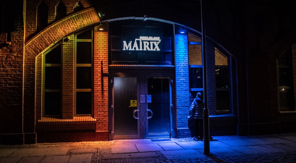 Drehorte Berlin Tag und Nacht Disco Matrix Eingang des Clubs „Matrix“ in Berlin bei Nacht. Die Backsteinfassade wird blau und orange beleuchtet, vor der Tür steht eine Person.