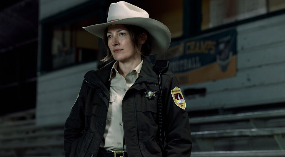 Auf dem Bild zur Lanterns-Serie steht eine Sheriff-Figur, gespielt von Kelly Macdonald, in einer nächtlichen Szene in einer Sporthalle oder Arena. Sie trägt einen weißen Cowboyhut, eine Sheriff-Uniform mit Abzeichen sowie eine dunkle Jacke und wirkt aufmerksam und entschlossen. Die unbekannte Sheriff-Figur blickt konzentriert zur Seite, während im Hintergrund Tribünen und ein unscharfes Hallenschild zu sehen sind.