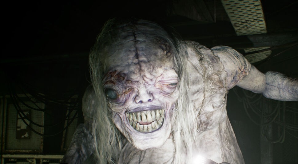 Auf dem Bild zu Resident Evil Requiem Tipps ist eine furchterregende Kreatur in einem düsteren, technisch wirkenden Raum zu sehen. Das Wesen hat eine bleiche, entstellte Haut, ein deformiertes Gesicht mit einem milchig-trüben Auge und ein unnatürlich breites, zahnreiches Grinsen. Lange, strähnige Haare fallen über seinen Körper. Die Szene wirkt beklemmend, bedrohlich und intensiv, typisch für die albtraumhafte Horroratmosphäre des Spiels.