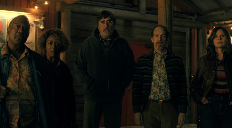 Auf dem Bild zu The Boroughs stehen Art, gespielt von Clarke Peters, Judy, dargestellt von Alfre Woodard, Sam, gespielt von Alfred Molina, Wally, gespielt von Denis O’Hare, und Renee, dargestellt von Geena Davis, nebeneinander vor einem hölzernen Gebäude. Alle blicken überrascht und angespannt nach vorne. Die warme Beleuchtung und ihre ernsten Gesichtsausdrücke unterstreichen die dramatische und geheimnisvolle Stimmung dieser Szene aus The Boroughs.