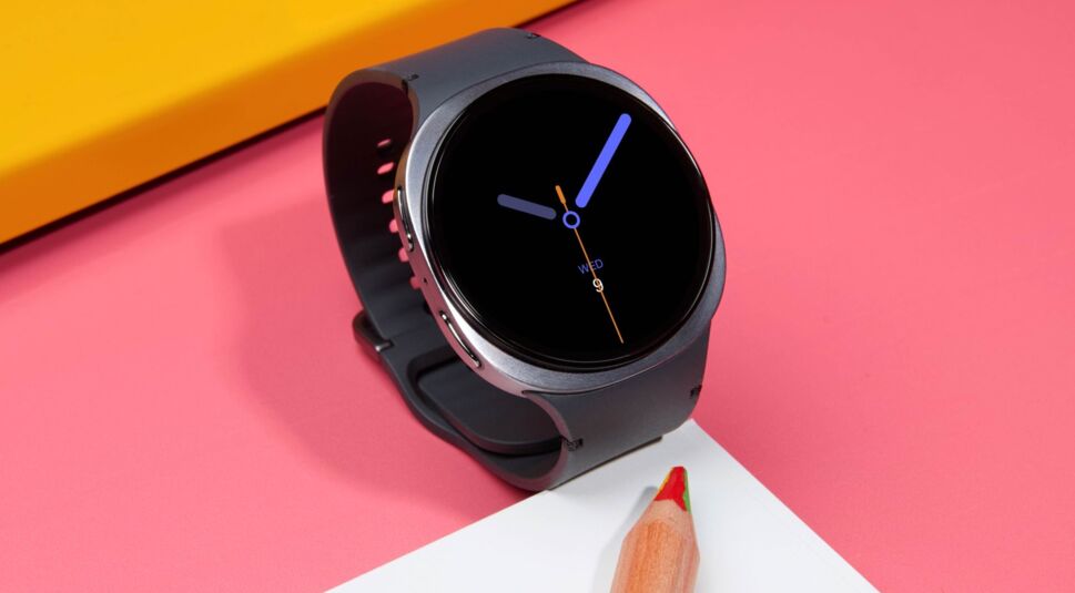Die Galaxy Watch8 in der Farbe Graphit liegt auf einem pinken Schreibtisch neben Papier und Stift.