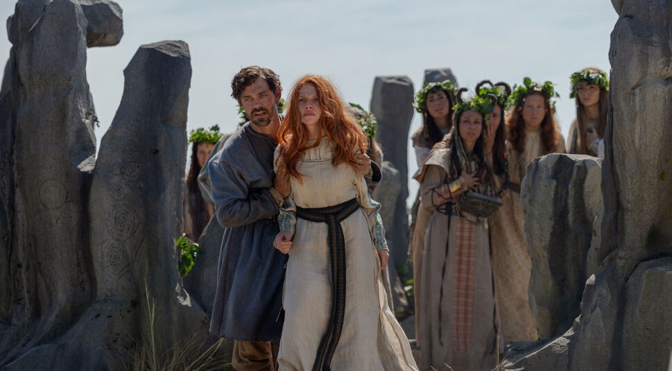 Auf dem Bild zu den GigaTV Highlights im Mai 2026 aus Der Medicus 2 sind Rob Cole, gespielt von Tom Payne, und Ilene, dargestellt von Áine Rose Daly, in einer dramatischen Szene zu sehen. Rob Cole steht hinter Ilene und hält sie schützend fest, während sie angespannt nach vorn blickt. Im Hintergrund stehen mehrere Frauen mit Kränzen, umgeben von großen Steinen, was eine mystische und bedrohliche Stimmung erzeugt.