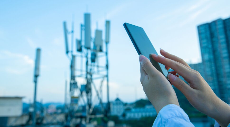 Auf dem Bild zum Xiaomi 5G Handy ist ein modernes Smartphone in der Hand einer Person zu sehen, die es im Freien benutzt. Im Hintergrund befinden sich unscharf mehrere Mobilfunkmasten sowie Gebäude, was auf eine urbane Umgebung hindeutet. Die Szene vermittelt eine technisch fortschrittliche und vernetzte Stimmung.