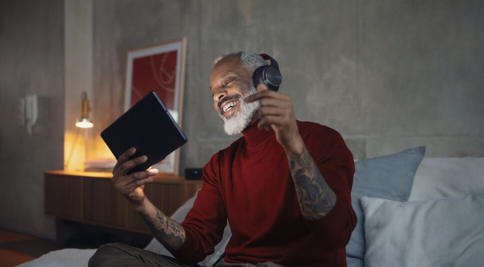 Auf dem Bild zu Google One Vodafone sitzt ein älterer Mann mit grauem Bart entspannt auf einem Bett und trägt kabellose Kopfhörer. Er hält ein Tablet in der Hand und lacht begeistert, während er Musik oder Inhalte streamt. Der Mann trägt einen roten Pullover und wirkt ausgelassen und zufrieden. Die warme Beleuchtung und das moderne Schlafzimmer schaffen eine gemütliche und entspannte Atmosphäre.