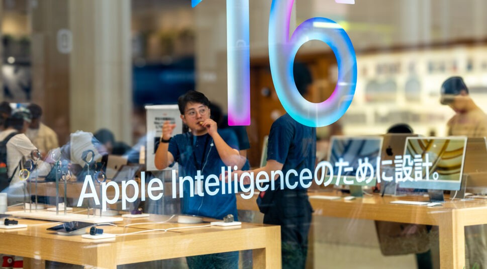 Auf dem Werbebild zu Apple Intelligence ist die Glasfront eines Apple Stores zu sehen, auf der groß der Schriftzug „Apple Intelligence“ steht. Im Hintergrund erkennt man Mitarbeiter in blauen Shirts sowie ausgestellte Geräte auf hellen Holztischen. Die Szene wirkt modern, geschäftig und technologisch fokussiert.