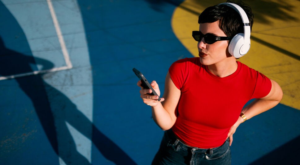 Auf dem Bild zum Thema Datenvolumen sparen steht eine Person mit kurzen dunklen Haaren, Sonnenbrille und weißen Kopfhörern auf einem farbigen Boden in Blau und Gelb. Sie trägt ein rotes Shirt, hält ein Smartphone in der Hand und wirkt konzentriert. Die starken Schatten und kräftigen Farben erzeugen eine moderne, dynamische Stimmung.