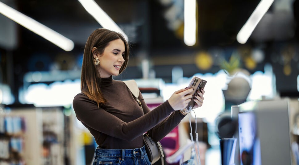 Auf dem Bild zum Handy-Speicher hält eine junge Frau mit langen braunen Haaren und braunem Rollkragenpullover ein Smartphone mit beiden Händen vor sich und betrachtet lächelnd das Display. Sie steht in einem hell beleuchteten Elektronikmarkt, im unscharfen Hintergrund sind Regale und Lichtbänder zu erkennen. Die Szene wirkt neugierig, entspannt und voller Vorfreude.
