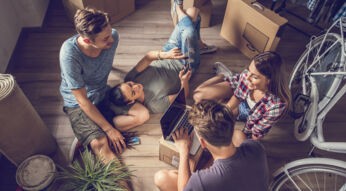 Vier junge Leute, die sich über WG-Portale gefunden haben, sitzen auf dem Boden zwischen Umzugskartons