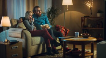 In the Evening Man and Woman are Sitting on the Sofa Watching TV and Eating Popcorn. Living Room is Cozy.