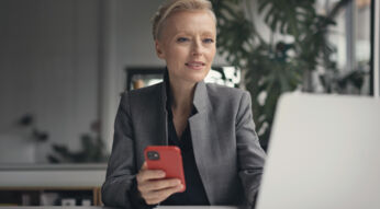 Eine Frau sitzt mit dem Smartphone in der Hand vor ihrem Laptop und prüft ihre Mobilfunk-Rechnung