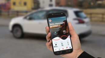 Hand hält Smartphone mit App von Vodafone und EMIL im Hintergrund steht ein weißes Auto.