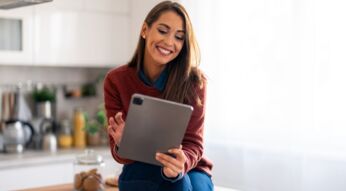 Lächelnde Frau mit iPad Pro sitzt auf dem Küchentisch.