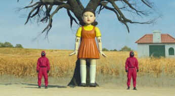 Zwei Wächter aus der Serie "Squid Game" stehen neben einer überdimensional großen Puppe.