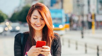 Junge Frau mit roten Haaren schaut lächelns auf ihr Smartphone.