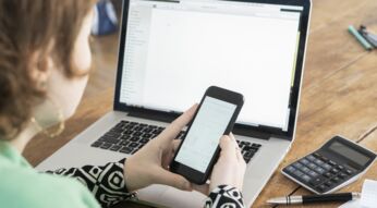 Frau sitzt vor Laptop und hält Smartphone in beiden Händen