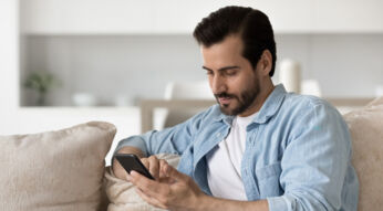 Mann mit Bart und blauem Hemd sitzt mit seinem Smartphone auf der Couch.