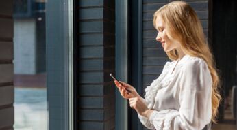 Junge Frau vor Fenster blickt lächelnd auf ihr Smartphone
