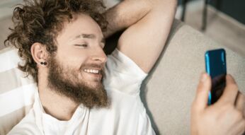Junger Mann mit Bart und verwuschelten Haaren liegt auf der Couch und guckt lächelnd auf sein Smartphone