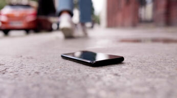 Auf dem Bild zum Thema Handy verloren oder gestohlen ist ein schwarzes Smartphone zu sehen, das unbeachtet auf einem grauen Gehweg liegt. Im unscharfen Hintergrund entfernt sich eine Person in Jeans und weißen Sneakern, links parkt ein rotes Auto, rechts ragt eine Backsteinfassade auf. Die bodennahe Perspektive und das gedämpfte Tageslicht vermitteln ein Gefühl von Verlust und Unachtsamkeit.