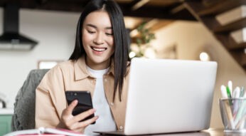 Lächelnde Frau sitzt vor Notebook und blickt auf Handy