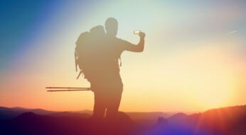 Das Foto zeigt einen Menschen mit Rucksack und Nordic-Walking-Stöcken, der ein Smartphone in die Luft hält