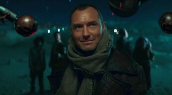 Jude Law als Jod Na Nawood in der Serie Skeleton Crew steht im Fokus des Bildes. Er trägt einen dicken Schal und eine dunkle Lederjacke, während er mit einem leichten Lächeln in einer futuristischen, dunkel beleuchteten Umgebung steht. Im Hintergrund sind unscharfe Gestalten und leuchtende Lichter zu erkennen, die eine mysteriöse Stimmung schaffen.