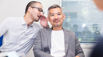 Das Bild zeigt zwei Männer. Der linke Mann flüstert dem rechten Mann etwas ins Ohr.